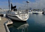 White sailboat moored at a wooden dock in a marina; calm blue water reflects the hull, with other boats in the background.