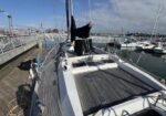 Sailboat docked at a marina with a wooden deck, mast, and gear secured onboard, calm water and other boats in the background