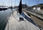 Sailboat tied at a marina, looking toward the bow along a wooden deck with a cabin hatch and rigging, harbor buildings in the background.