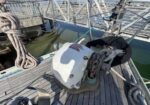 White anchor windlass on a wooden sailboat deck with coiled rope and rigging; water visible beyond the railing.