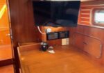 Wood-paneled boat cabin with a wall-mounted flat-screen TV, small electronics, and a desk area; window to the right.