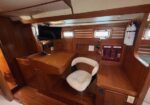 Wood-paneled boat cabin navigation/chart station with a curved desk, white cushioned chair, mounted monitor, and a red switch panel; fire extinguisher on the wall.