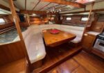 Wood-paneled sailboat cabin with a U-shaped white sofa around a wooden dining table and a compact galley on the right.