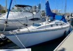 White sailboat moored at a marina, with blue sail cover and rigging; city skyline in the background.