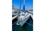 White sailboat moored in a marina, bow facing forward, with a blue sail cover and rigging, surrounded by other boats.