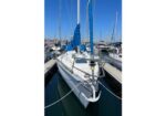 White sailboat docked in a marina, with blue sail covers and surrounding boats in the harbor