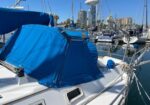 Sailboat in a marina with a bright blue dodger and cockpit cover, masts and rigging, blue sky, and city buildings in the background.