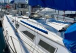 White sailboat docked in a marina with blue cover and windows along the hull; other boats visible in the background.