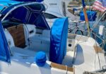 White sailboat cockpit with blue canvas covers, open hatch, and wooden accents, docked at a marina with an American flag nearby.