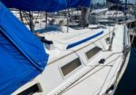 White sailboat deck with blue canvas cover and rigging, moored among other boats in a marina.