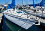 Sailing yacht docked in a sunny marina with a white hull, blue covers, and surrounding sailboats.