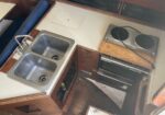 Compact boat galley with a double stainless steel sink, wooden cabinets, and a two-burner stove/oven unit beneath.