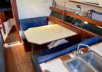 Interior of a sailboat cabin showing a dinette with an octagonal table and blue cushioned benches, wood trim, and a double sink nearby on the galley counter