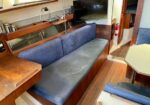 Inside a boat cabin: blue cushioned bench along the wall, wooden table, mounted TV, and a doorway with a fire extinguisher nearby.