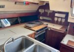 Compact boat galley with a stainless sink, two-burner stove on a wooden counter, and an oven beneath the counter. Built-in shelves and curved wall panels surround the area.