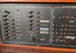 Close-up of a vintage boat dashboard with two panels: left 12 VDC accessory controls (DC MAIN, cabin lights, running lights, water pump, depth sounder, autopilot) and right 120 VAC load center with switches and indicator lights.
