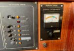 Close-up of a wooden boat electrical panel showing a 120 VAC load center with switches and a professional electrical system monitor gauge.