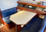 Boat cabin dining area with blue cushioned benches and a light hexagonal table; a small hatch sits in the center of the table.