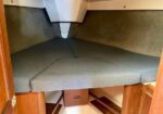 Interior of a small boat cabin with a V-shaped cushioned bed in dark gray fabric, wooden cabinetry and storage underneath.