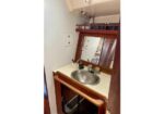 Compact boat galley sink area: round stainless sink in beige countertop, wood cabinet below, mirror and shelf above, with an electrical outlet on the wall.