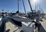 Sailboat with white hull docked at a sunny marina, with masts of other boats rising along the pier and blue sky overhead.
