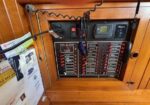 Wooden cabinet housing a boat electrical panel with labeled toggle switches and a digital battery monitor