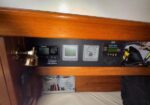 Boat cabin electrical panel with multiple digital gauges and a brass reading lamp under a wooden shelf above a bed.