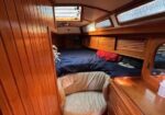 Cozy wooden boat cabin with built-in bunks along both sides, navy blankets, and small porthole windows above; wood-paneled walls and a dresser visible.