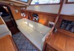Inside a wooden yacht cabin: cream cushioned bench seat along a panel-lined wall, navigation desk with controls, framed photos, and sunlight streaming through porthole windows.