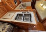 Wooden yacht galley with a double stainless-steel sink and beige countertops, seating area visible in the cabin.