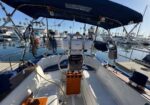 Cockpit of a sailboat docked at a marina, steering wheel centered, wooden pedestal, and blue canopy, with other boats in the background.