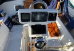 Helm of a sailboat showing three navigation displays and control instruments mounted on a pedestal, with sunlit wooden deck and equipment around.