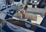 Sailboat cockpit with a wooden deck area, stainless steel rails, steering wheel, winch, and hatch, with a marina in the background.