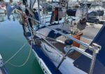 Blue and white sailboat moored in a marina, with stainless rails and lines securing it, surrounded by other boats on calm water.