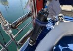 Close-up of a black cylindrical device strapped to a sailboat railing, with a gloved hand nearby and water in the background.