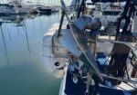 Stern of a sailboat with a metal anchor and docking gear, moored in a calm marina with other boats in the background.