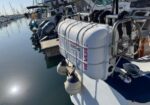 White propane tank mounted on a boat's stern, secured with a metal brace, with fenders hanging over the side and a marina in the background.
