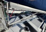 Close-up of a sailboat deck showing rigging, lines, and winches under a blue sail cover; metal supports and cockpit hatch are visible.