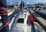 View from the cockpit of a white sailboat docked in a marina, with other boats, masts, and red life-raft containers along the deck.