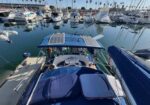 View from the stern of a blue boat with cushioned seating, solar panels on the canopy, docked among other boats in a marina.