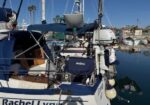 Sailboat docked in a marina, stern name 'Rachel Lynn' visible, with blue covers, rigging, fenders, and reflections in calm water.