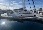 Sailboats docked at a marina with calm water reflecting hulls and tall masts under a sunny sky.