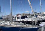 Blue sailboat in the foreground at a busy marina, with masts, rigging, and other yachts in the background under a clear blue sky.