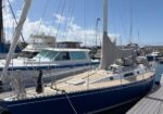 Blue sailboat docked at a marina, with a furled mainsail and beige deck, moored beside other boats under a blue, partly cloudy sky.