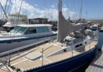Sailing yacht docked at a marina; beige deck, furled mainsail, and neighboring motorboats in the background.