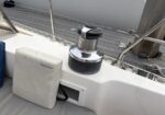 Sailboat deck with a chrome winch, black base, and a padded West Marine bag leaning against the cockpit wall; railing and dock in the background.