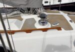 Close-up of a sailboat deck with winches, rope hardware, and a wooden handrail on a beige non-slip surface.