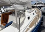 White sailboat docked at a marina with an open cockpit, wooden interior visible, and a blue hull beside a gangway.