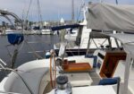 View from the deck of a sailboat in a marina, showing a wooden hatch, steering wheel, ropes, and surrounding docked boats with masts in the background.