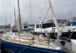 Blue sailboat docked at a marina, beige deck with rope rigging, surrounded by other moored boats on a cloudy day.